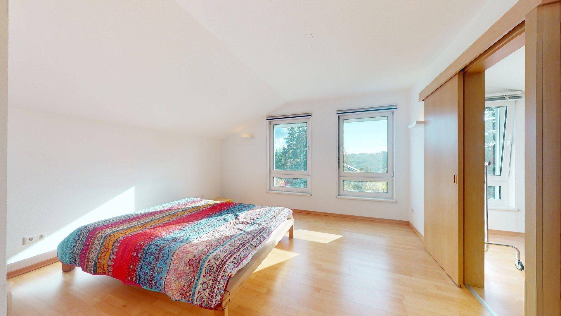 Schlafzimmer mit Holzboden, zwei Fenstern und einem Ausblick ins Grüne, unter einem Dachschrägenbereich.