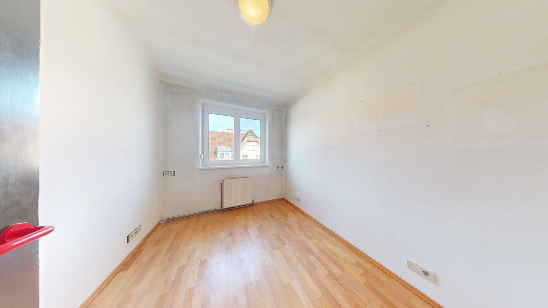 Helles, leeres Zimmer mit Parkettboden und einem Fenster, ideal als Schlafzimmer oder Arbeitszimmer nutzbar.