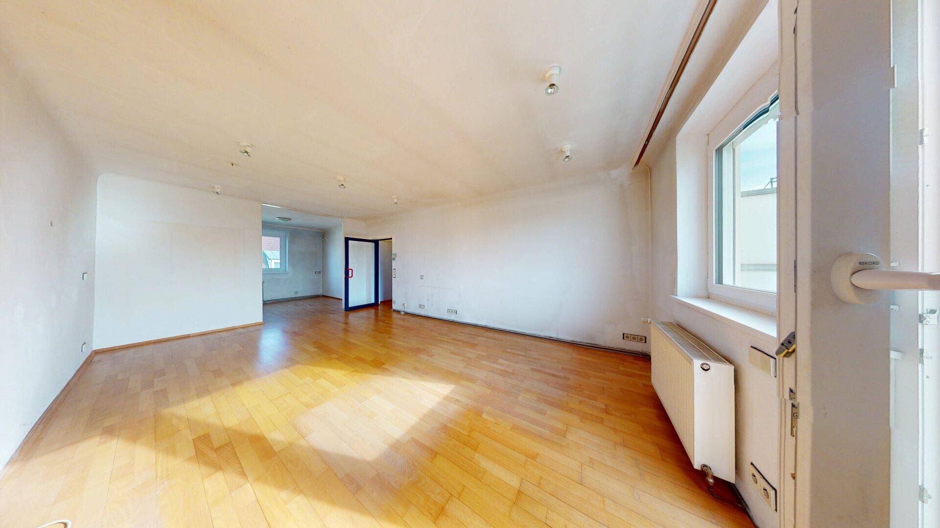 Geräumiges Wohnzimmer mit hellem Parkettboden und großen Fenstern, die viel Tageslicht hereinlassen.