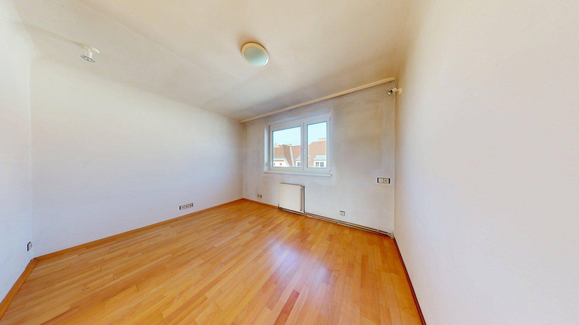 Helles Zimmer mit Parkettboden und einem Fenster, das einen Blick auf die umliegenden Dächer bietet.