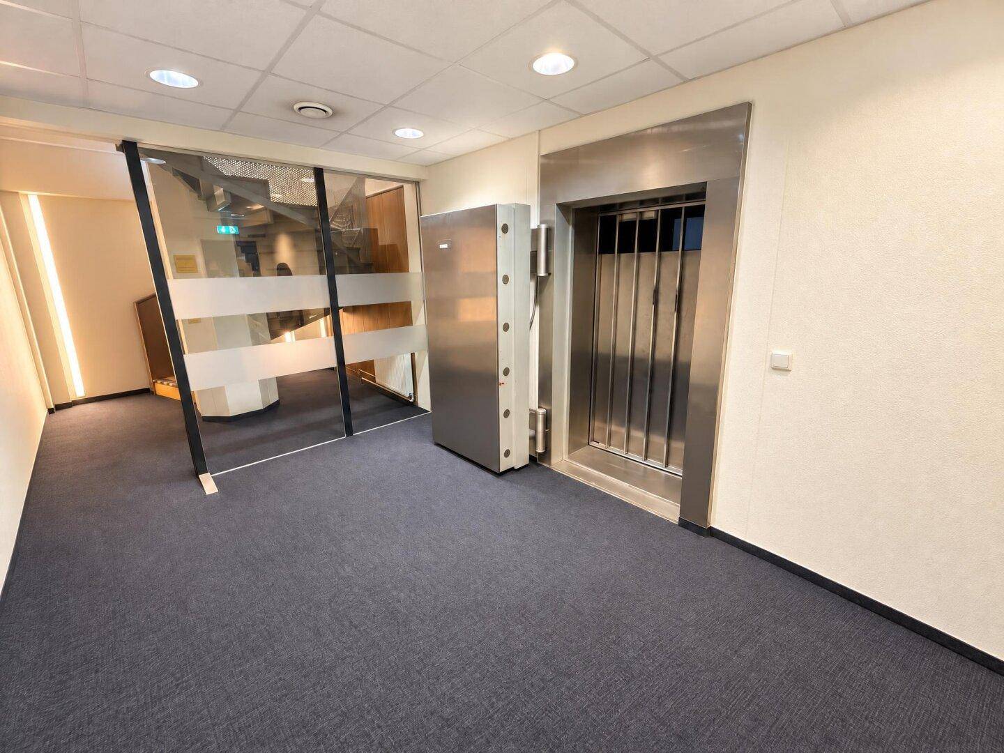 Heller Flur mit Teppichboden, Glasabtrennung und einem modernen Aufzug aus Edelstahl.