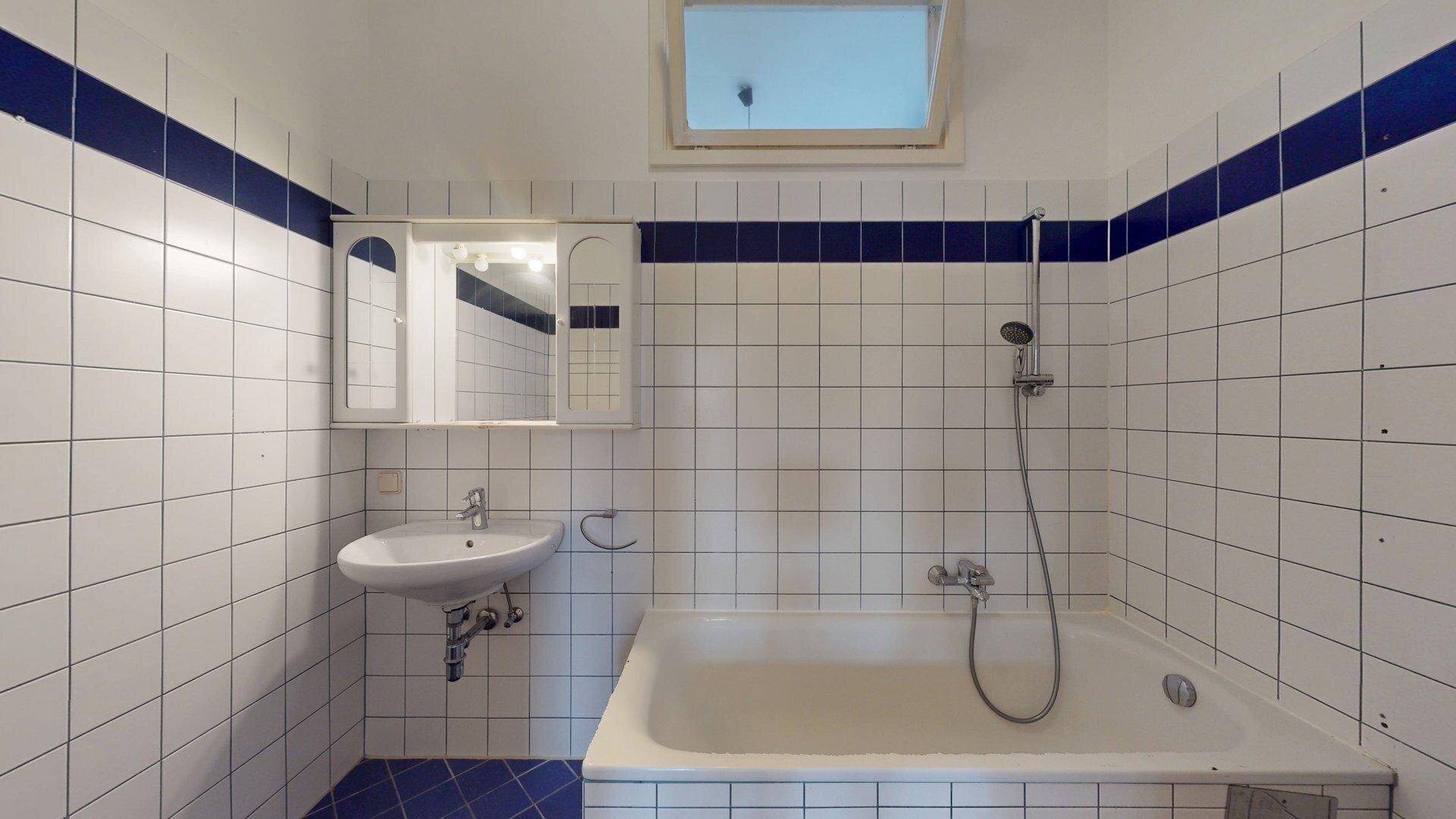 Badezimmer mit weißen Fliesen, blauen Akzentstreifen, Badewanne, Waschbecken und Fenster für Tageslicht.