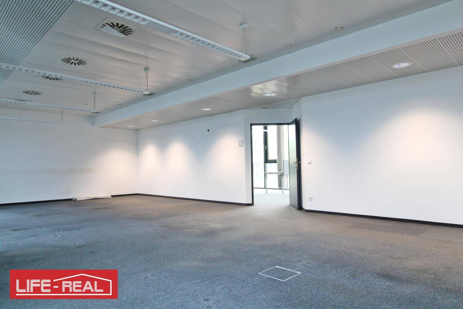 Leeres Büro mit Teppichboden, weißen Wänden und einer offenen Tür, die den Blick in einen weiteren Raum freigibt.