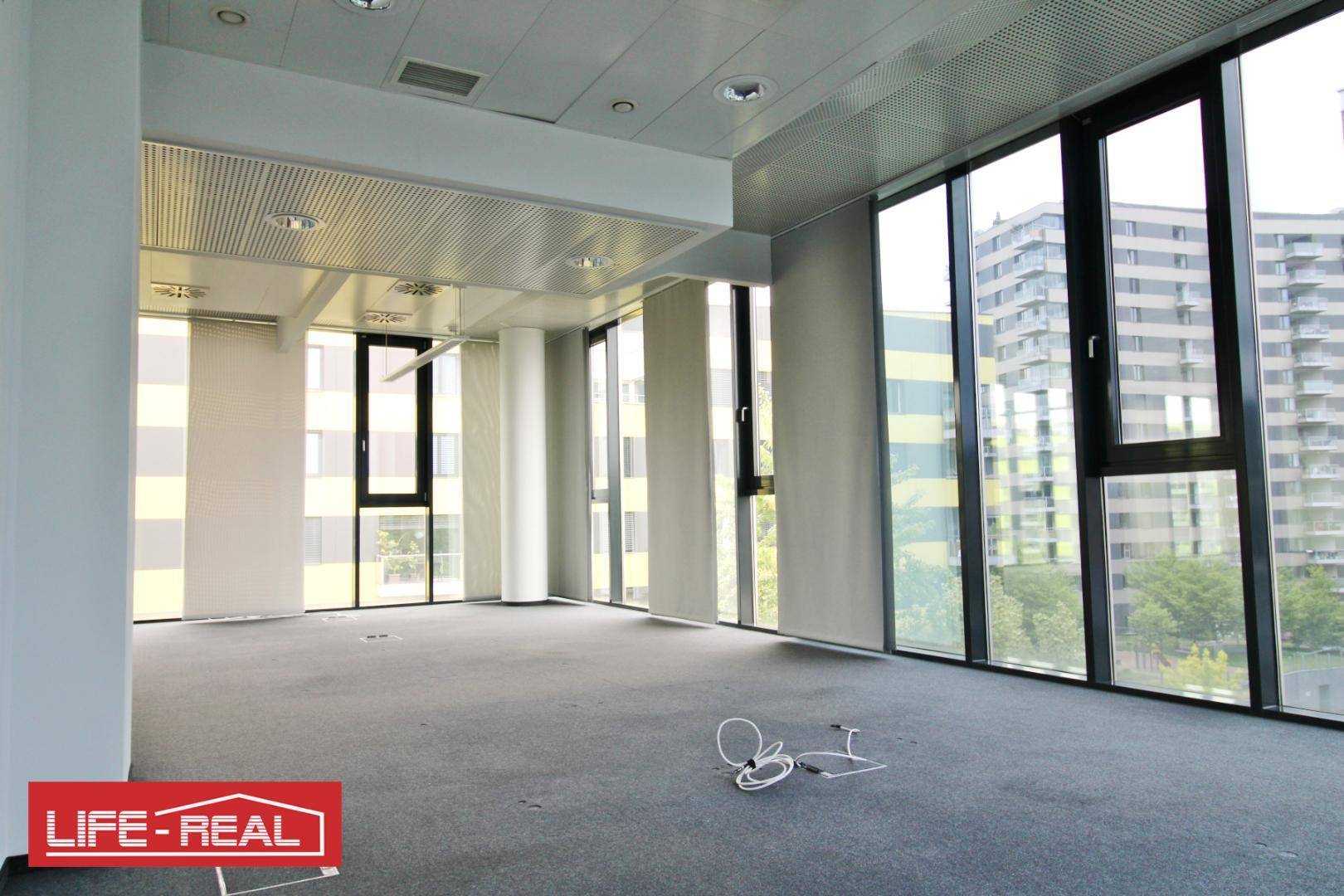 Geräumiges Büro mit Teppichboden und großen Fenstern, die einen weiten Blick auf die umliegenden Gebäude und die Stadt bieten.