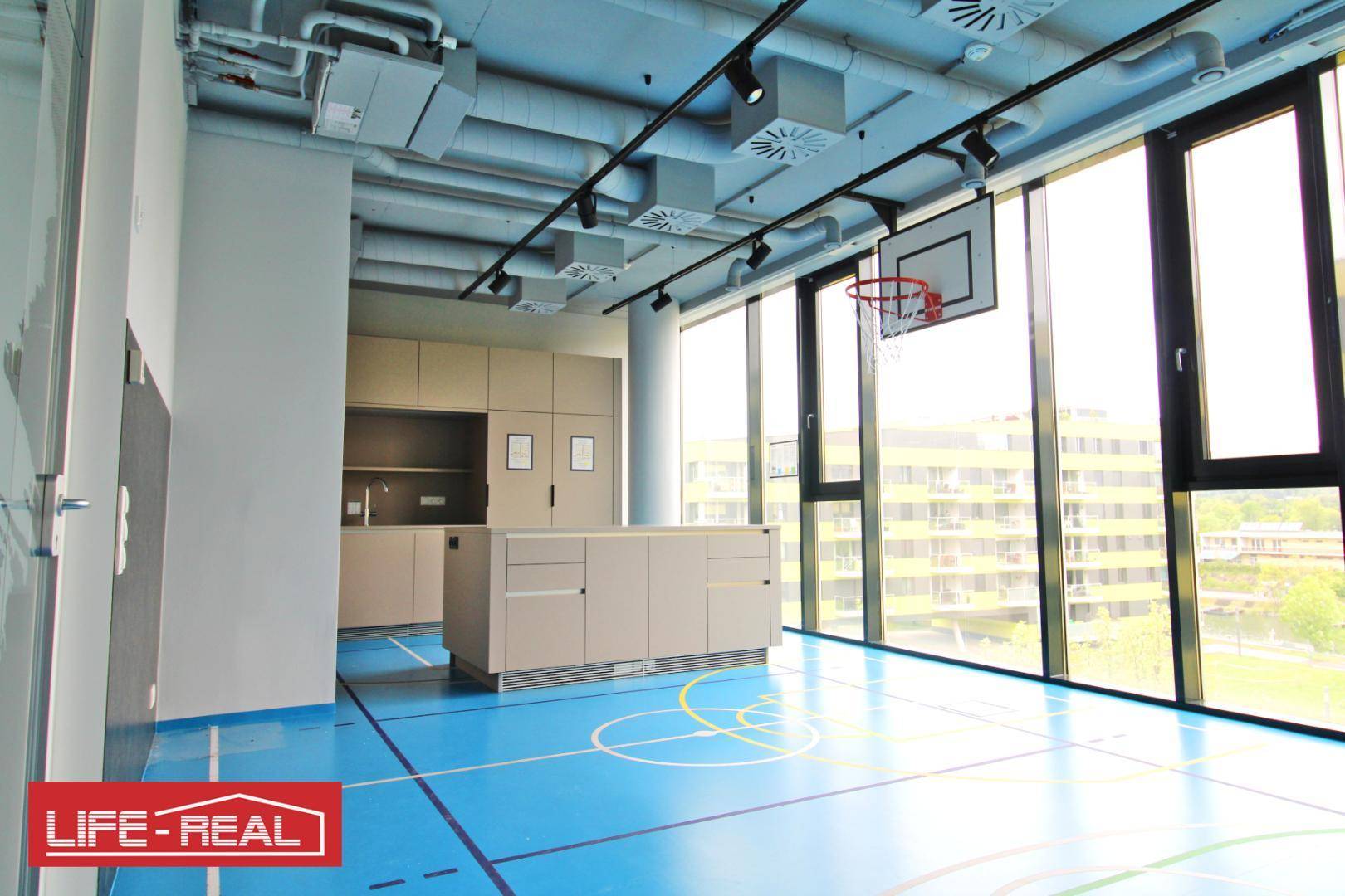 Vielseitiger Freizeitraum mit Basketballkorb, blauer Sportbodenmarkierung und integrierter Küchenzeile, ideal für Pausen.