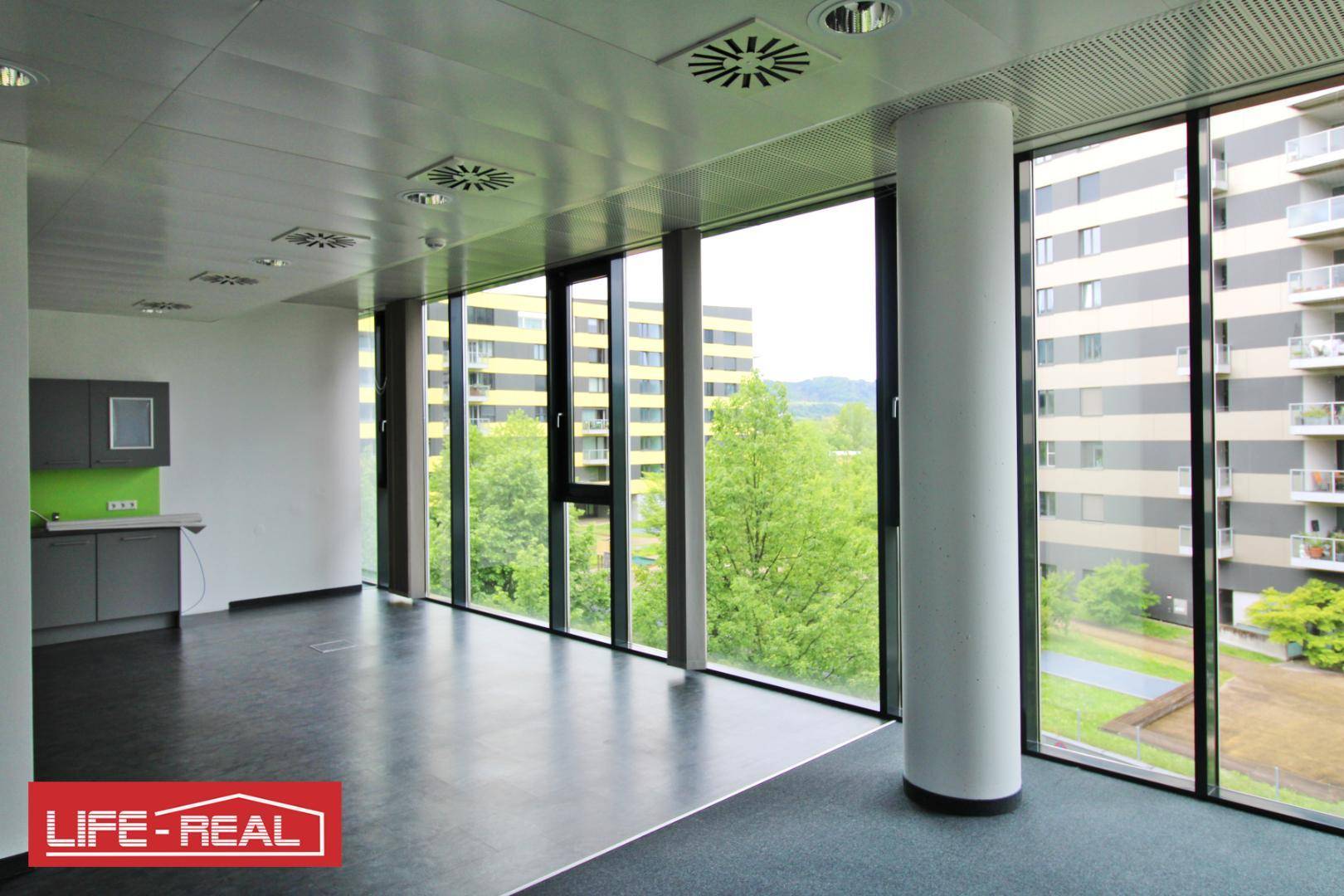 Büro mit integrierter Küchenzeile, großen Fenstern und Blick auf die umliegenden Gebäude und Grünflächen.