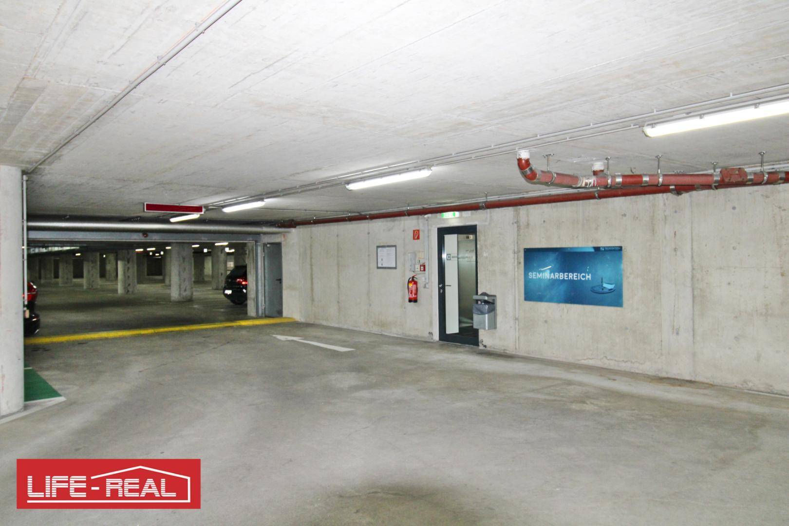 Tiefgarage mit Betonwänden, Deckenbeleuchtung und Hinweisschildern, die den Weg zu den Parkplätzen weisen.