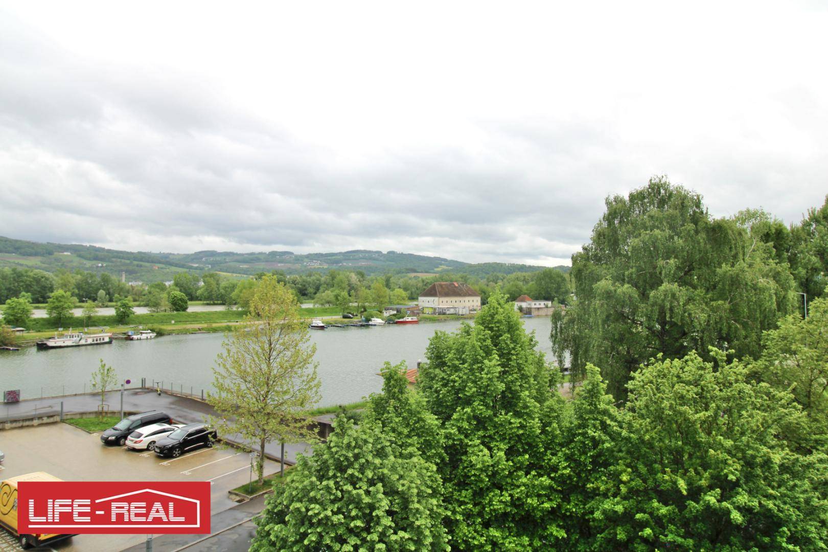 Panoramablick auf einen Fluss mit Booten, umgeben von grünen Bäumen und sanften Hügeln unter bewölktem Himmel.