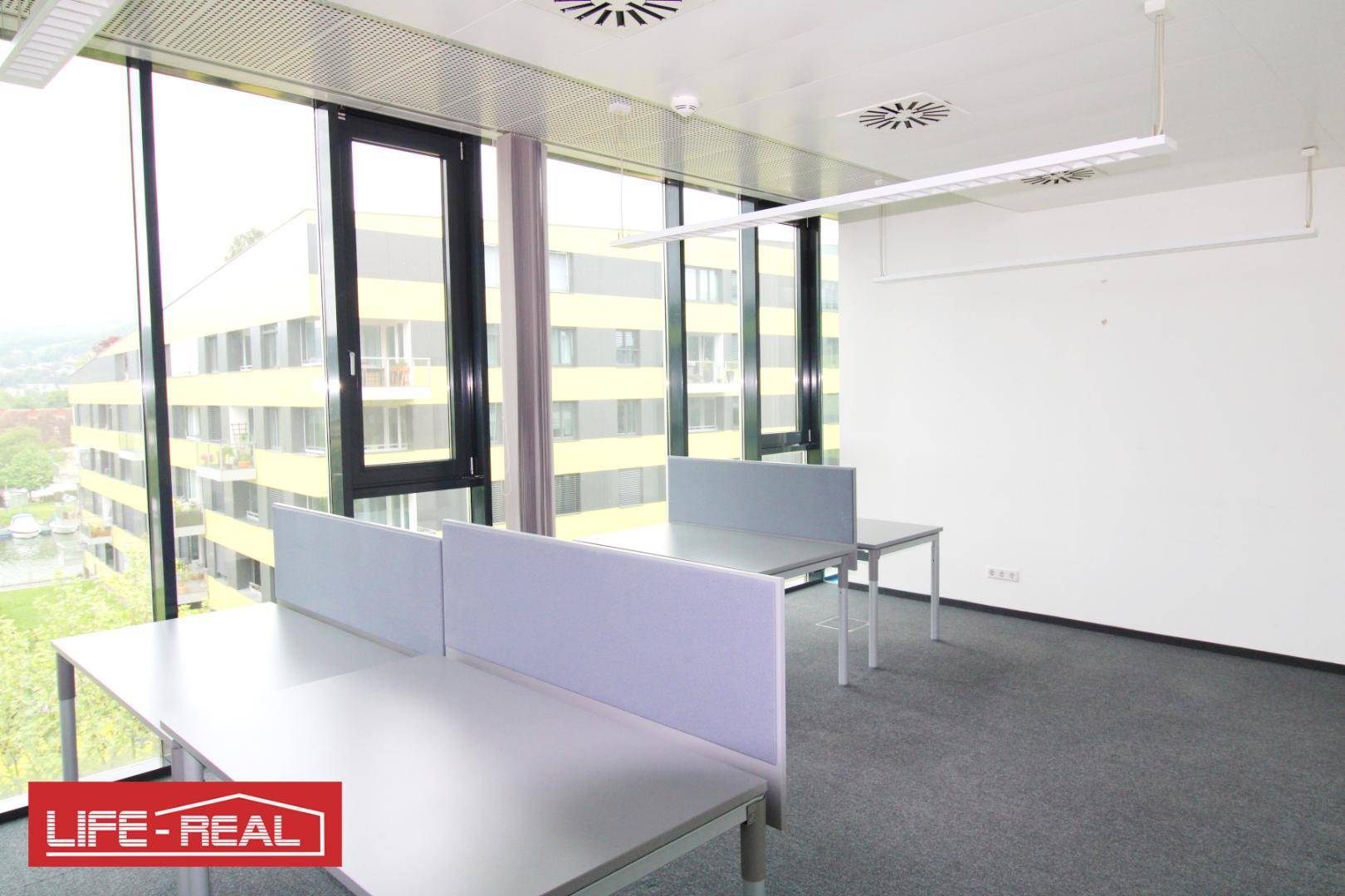 Büro mit mehreren Schreibtischen, großen Fenstern und Blick auf die moderne Architektur.
