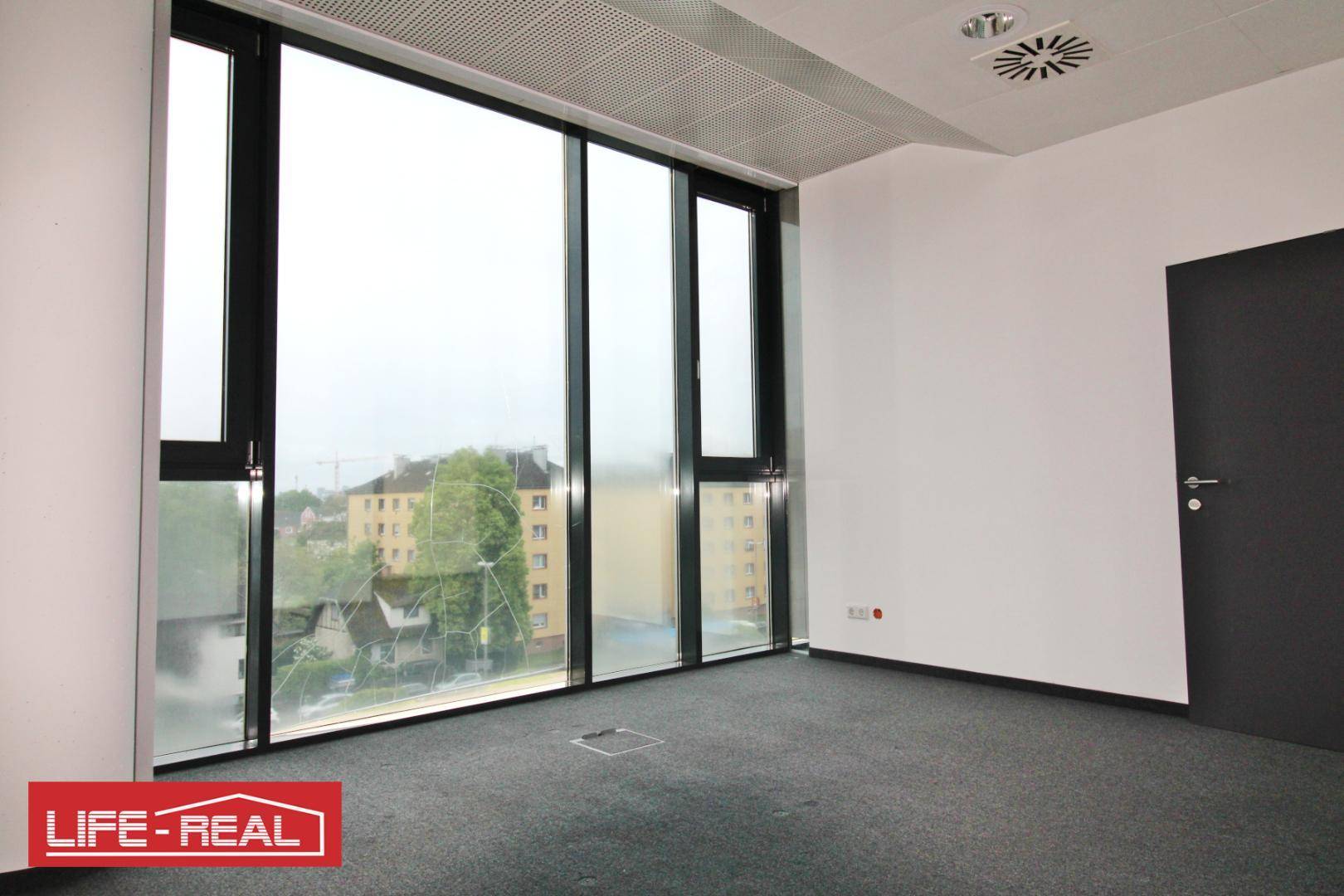 Helles Büro mit großen Fenstern, Teppichboden und Blick auf die städtische Umgebung.