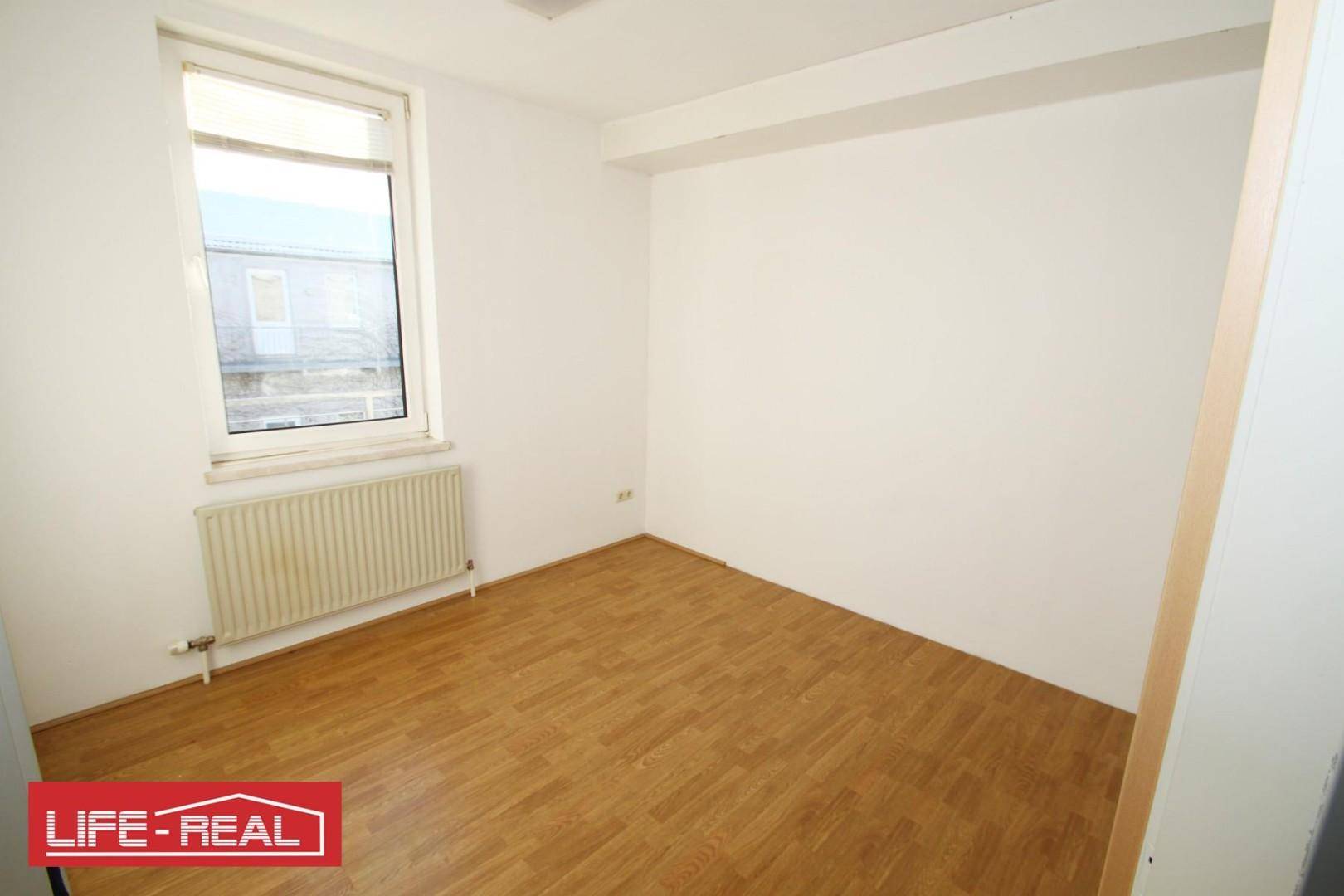 Leeres Zimmer mit Laminatboden, weiß gestrichenen Wänden und einem Fenster mit Heizkörper darunter.
