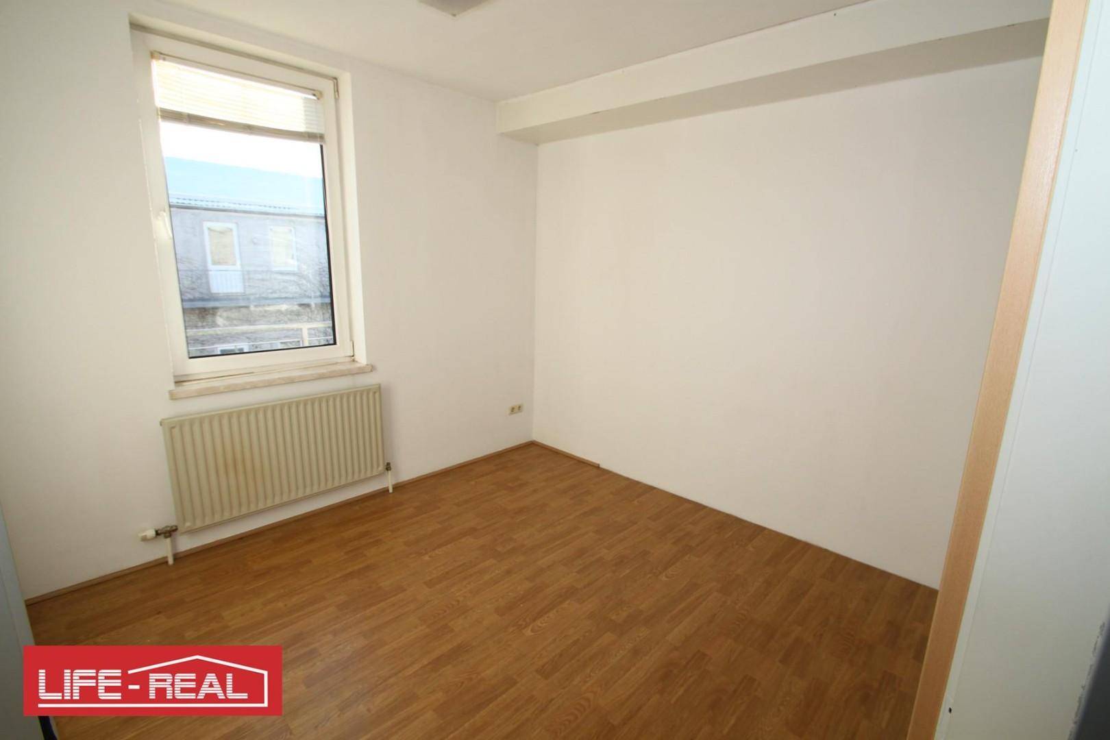 Leeres Zimmer mit Laminatboden, weiß gestrichenen Wänden und einem Fenster mit Heizkörper darunter.
