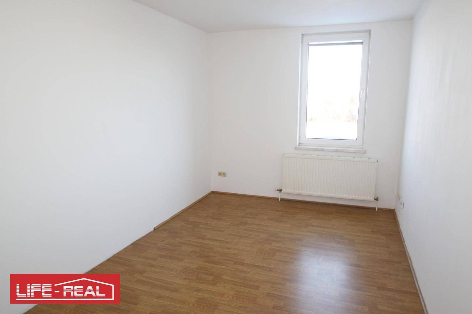 Helles, leeres Zimmer mit Laminatboden, weißen Wänden und einem Fenster, das viel Tageslicht hereinlässt.