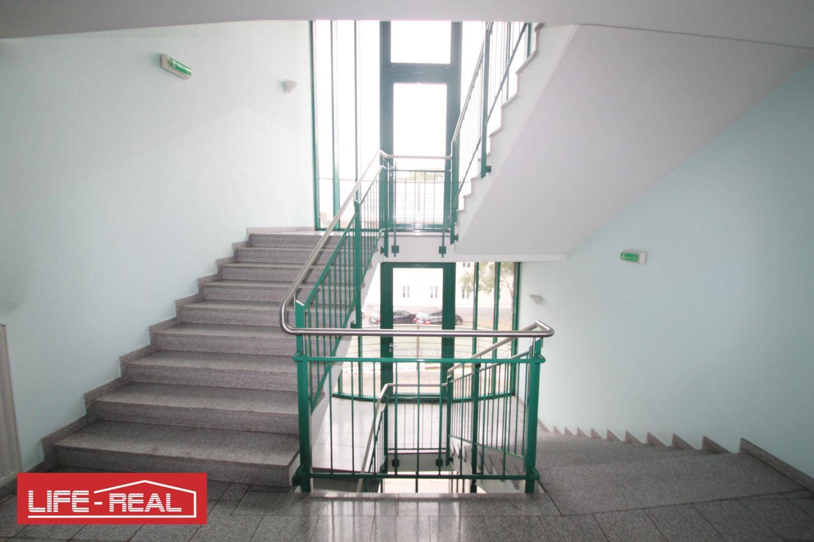 Geräumiges Treppenhaus mit grünem Geländer und großen Fenstern, das viel Tageslicht hereinlässt.