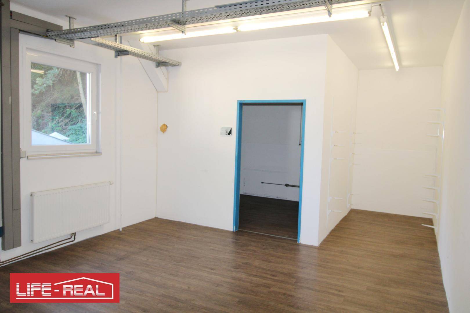 Heller Büroraum mit Fenster und Heizkörper, ausgestattet mit Laminatboden und blauer Tür.