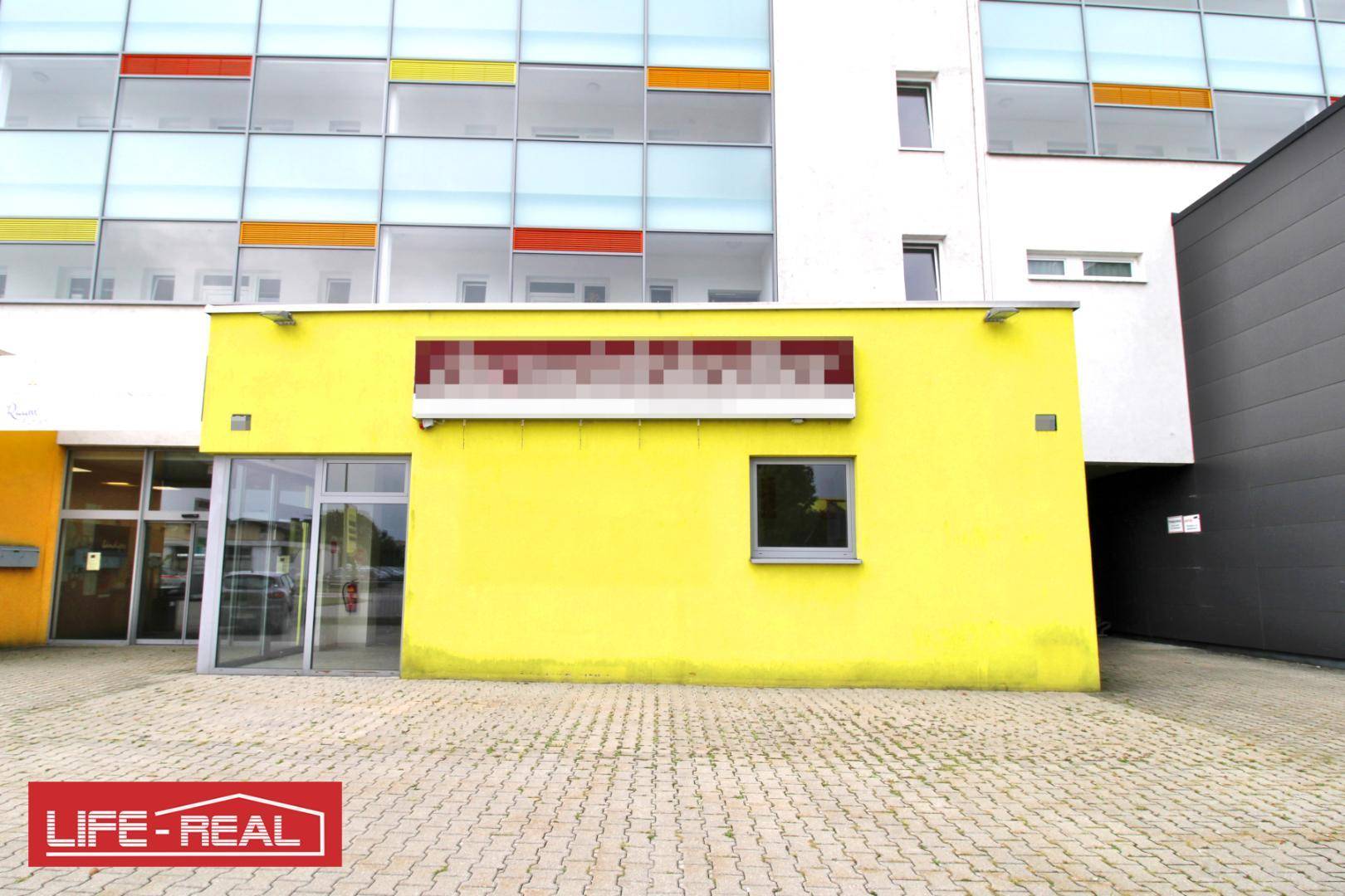 Moderne Gebäudefassade mit leuchtend gelbem Anbau und großen Fenstern, umgeben von einem gepflasterten Parkplatz.