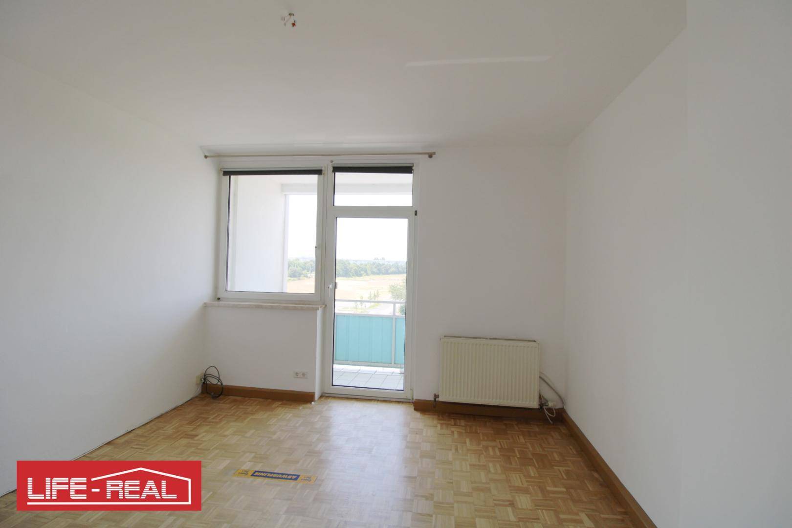 Leeres Zimmer mit Parkettboden und Zugang zu einem kleinen Balkon, bietet viel Tageslicht.