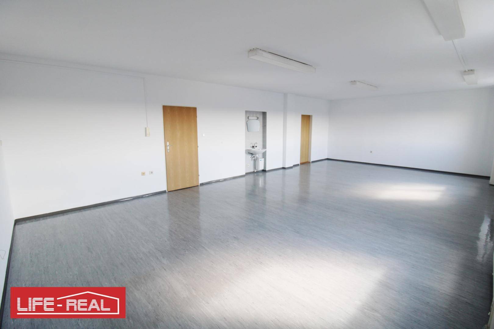 Großer, leerer Büroraum mit glänzendem Vinylboden, weißen Wänden und einem Waschbecken.