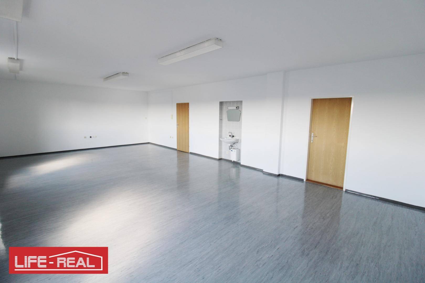 Geräumiger, heller Büroraum mit Vinylboden, weißen Wänden und mehreren Holztüren.