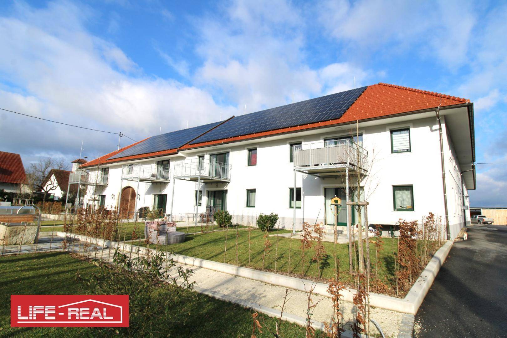 Modernes Gebäude mit Photovoltaikanlage auf dem Dach und Balkonen, umgeben von Grünflächen.