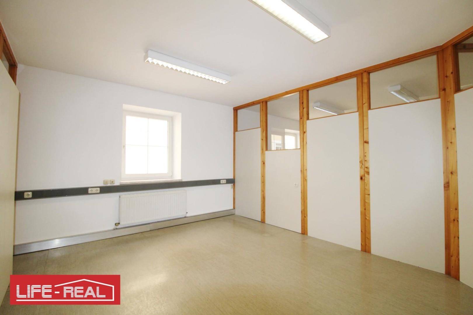 Heller Büroraum mit einem Fenster, Heizkörper und Holz-Glas-Trennwänden, die den Raum strukturieren.