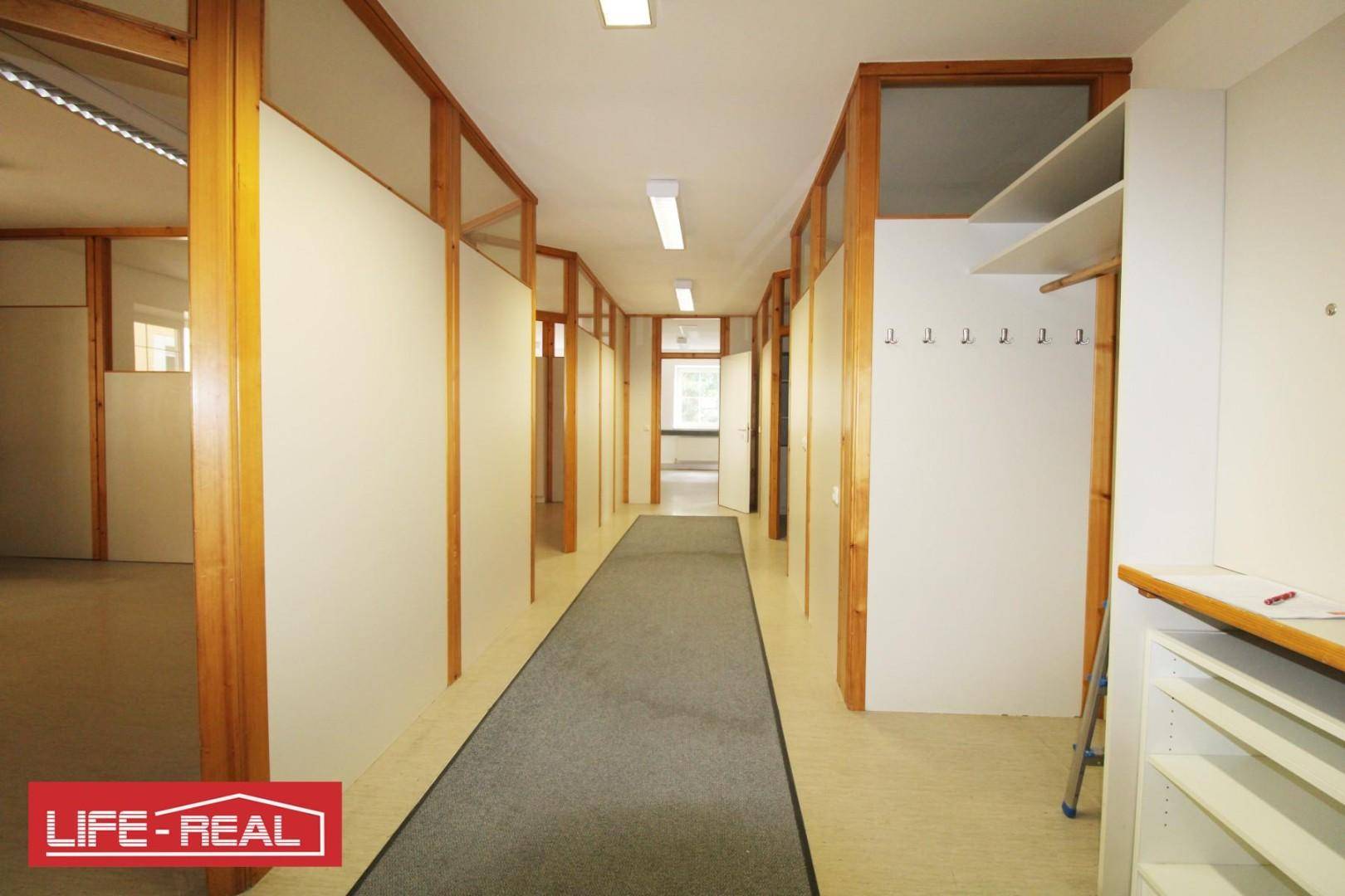 Langer Flur mit Holz-Glas-Trennwänden, Teppichboden und Garderobenbereich, der zu weiteren Büroräumen führt.