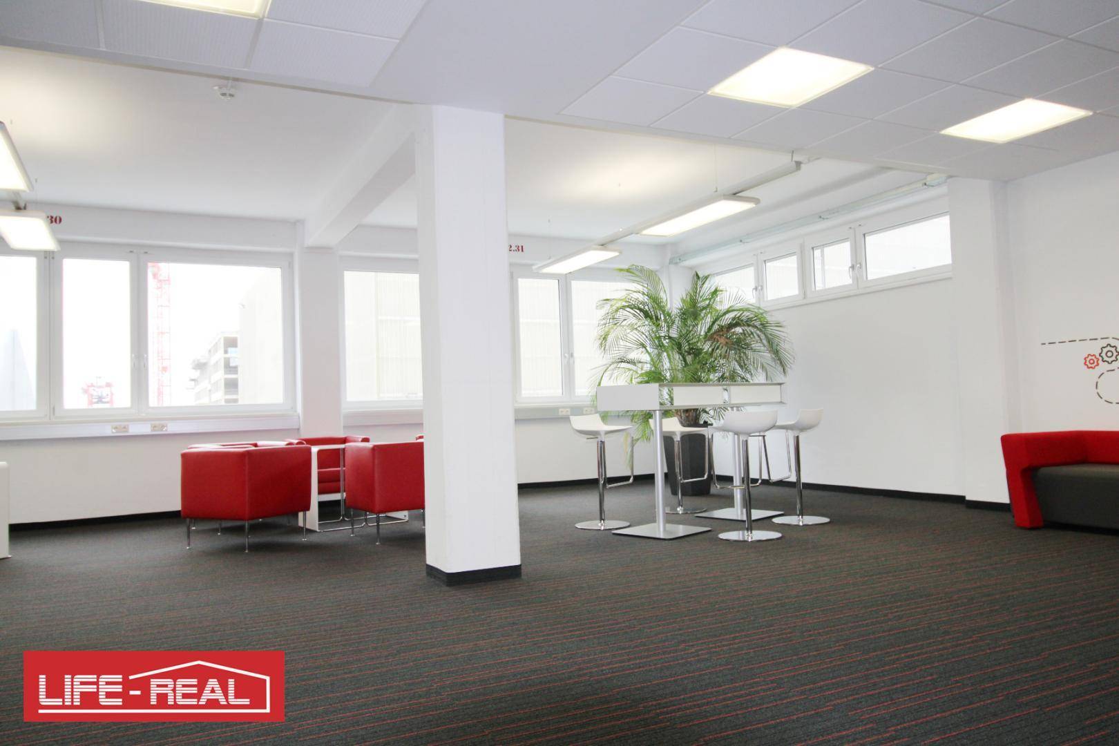 Heller Büroloungebereich mit roten Sitzmöbeln, Stehtischen und großen Fenstern, ideal für Pausen.