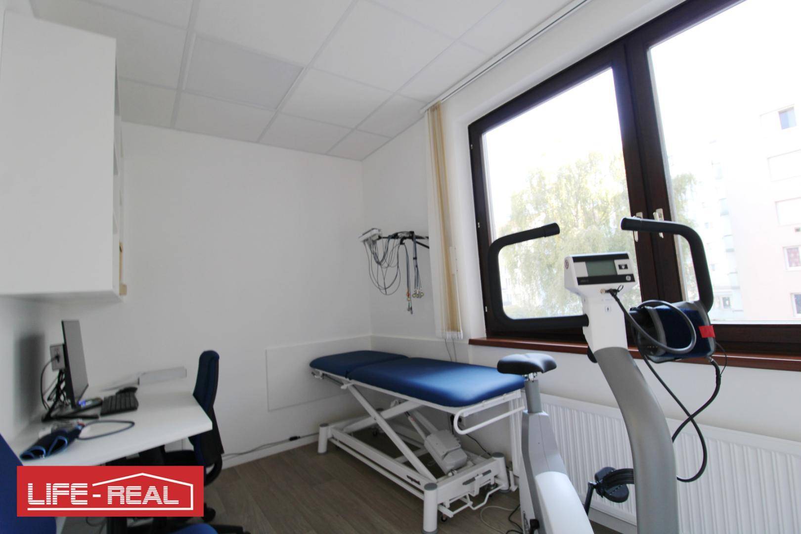 Heller Behandlungsraum mit medizinischem Ergometer, Untersuchungsliege und großem Fenster.
