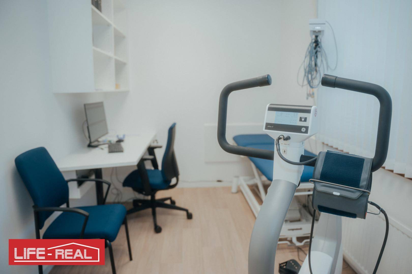 Behandlungsraum mit medizinischem Ergometer, Untersuchungsliege und Schreibtisch für Diagnostik.