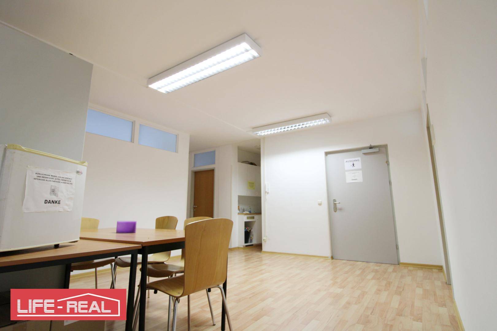Funktionaler Pausenraum mit Tisch, Stühlen und Kühlschrank, ideal für Mitarbeiter oder Besprechungen.