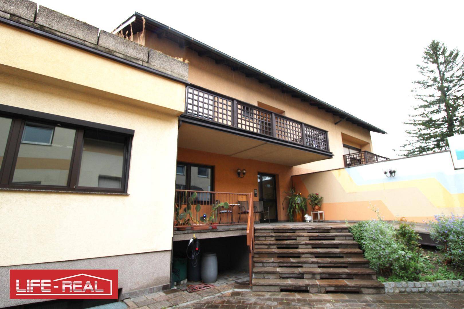 Außenansicht des Gebäudes mit Balkon, Außentreppe und umliegender Bepflanzung.