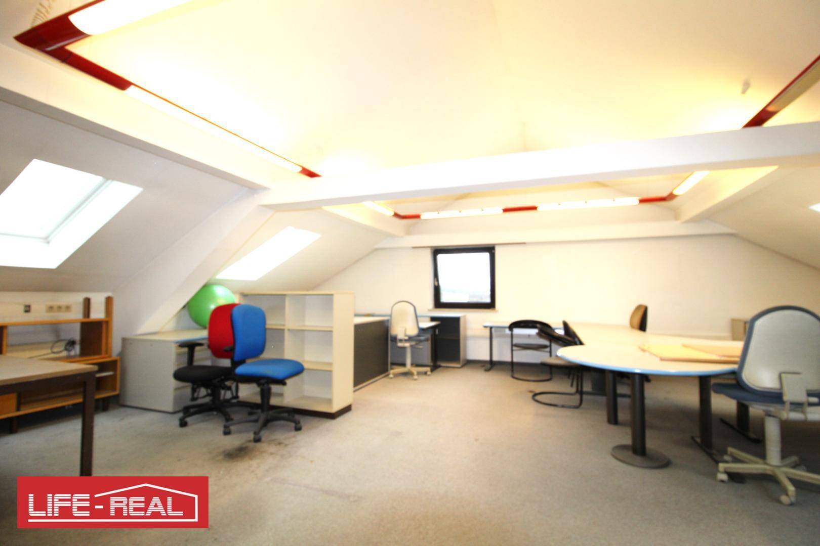 Helles Dachgeschossbüro mit mehreren Arbeitsplätzen, Regalen und schrägen Wänden, gut beleuchtet durch Dachfenster.