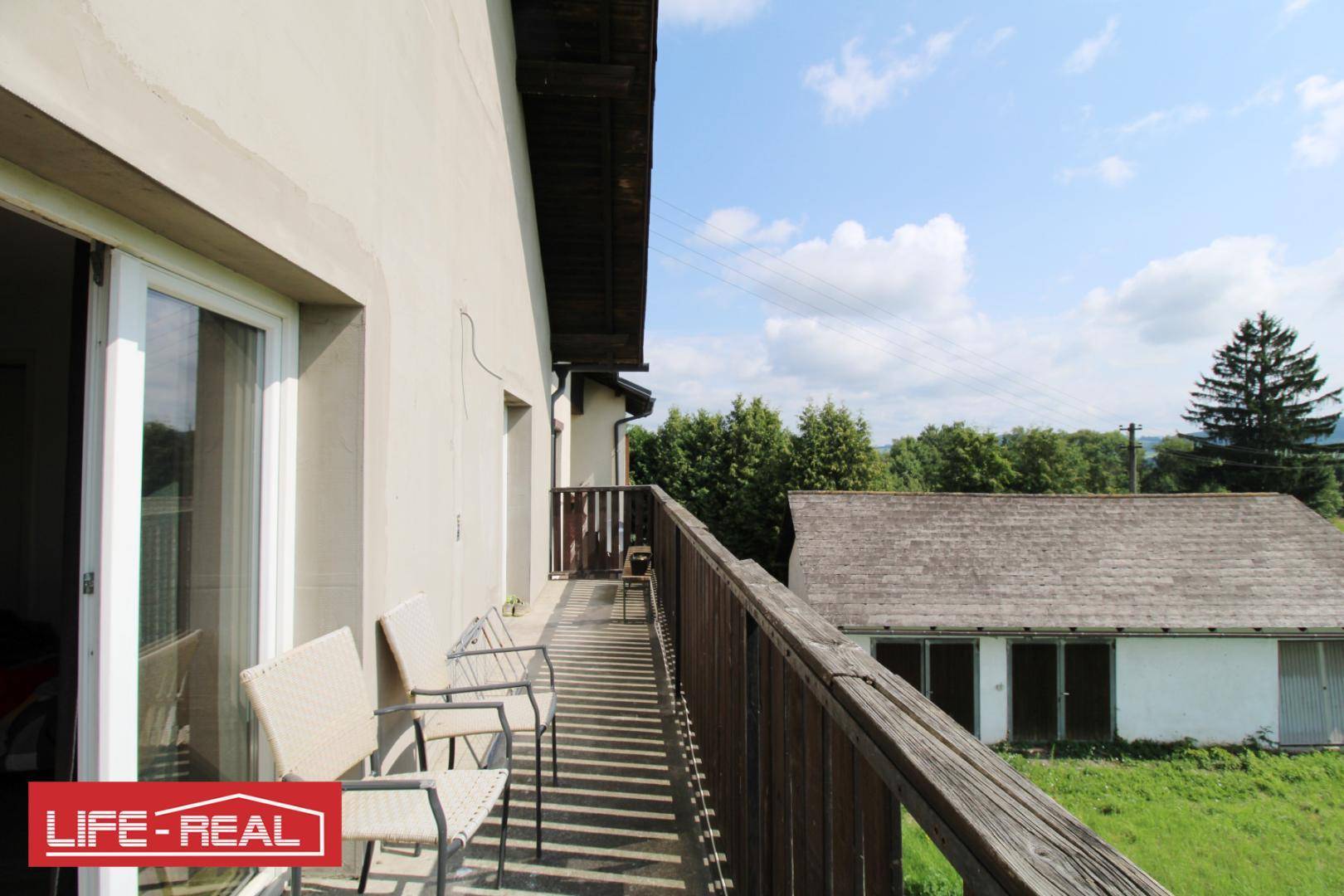 Balkon mit Sitzgelegenheiten und Blick auf ein Nebengebäude sowie weite grüne Felder und Landschaft.