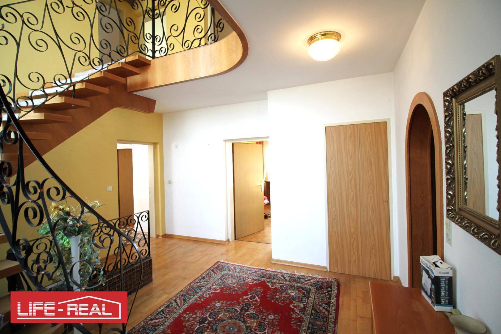 Heller Flur mit geschwungener Holztreppe, schmiedeeisernem Geländer und Parkettboden.