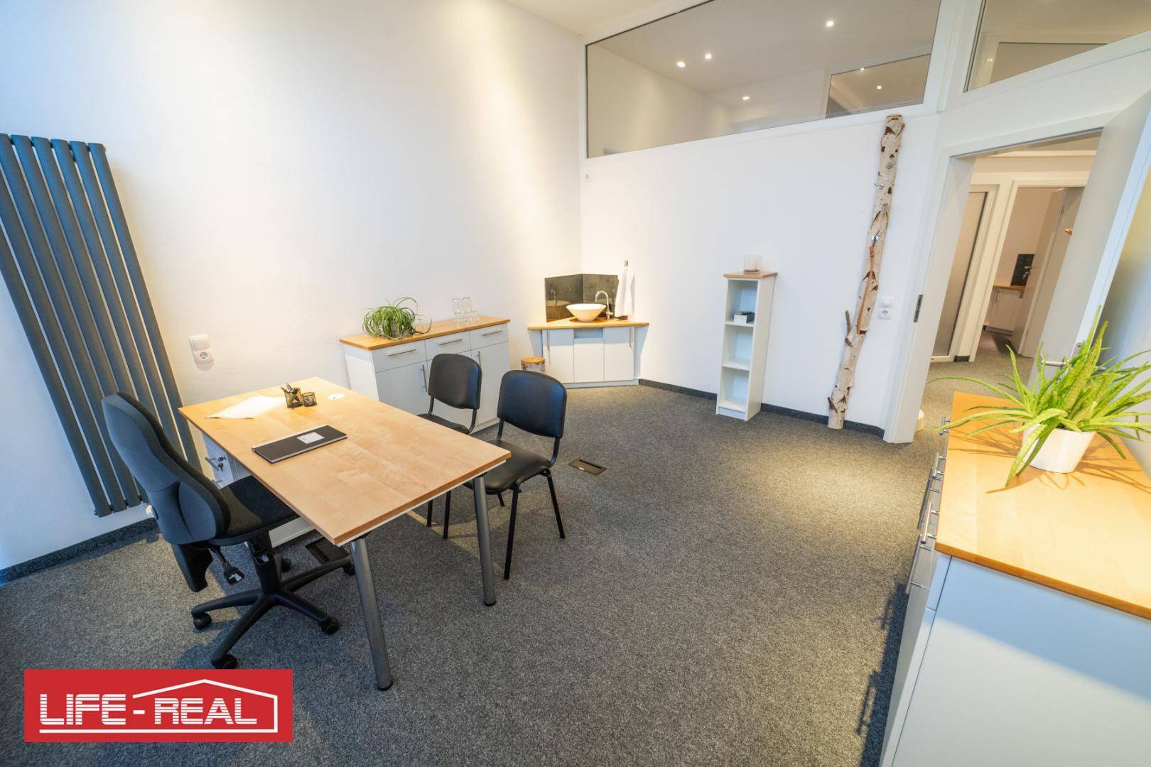 Geräumiger Büroraum mit Schreibtisch, Stühlen, Teppichboden und einem kleinen Waschbeckenbereich.