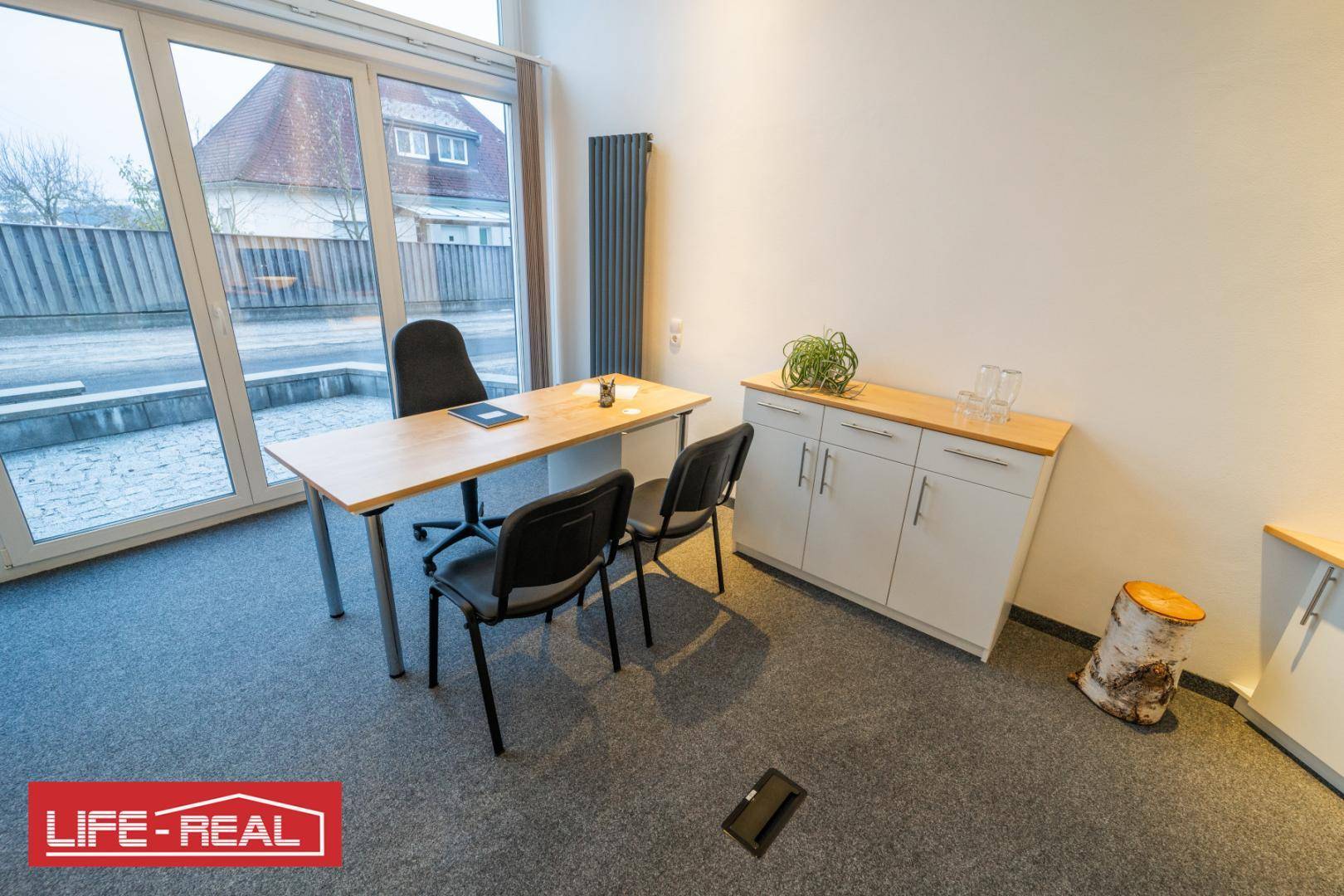 Heller Büroraum mit Schreibtisch, Stühlen, Teppichboden und großen Fenstern mit Blick nach draußen.