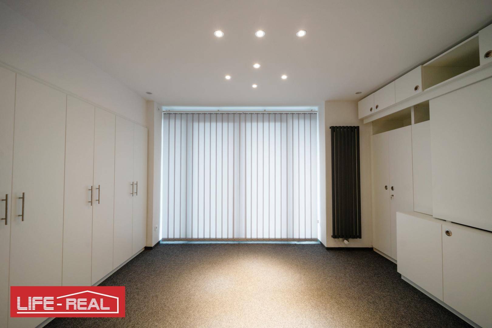 Geräumiger Raum mit Einbauschränken, Teppichboden, Deckenstrahlern und vertikalen Jalousien vor dem Fenster.