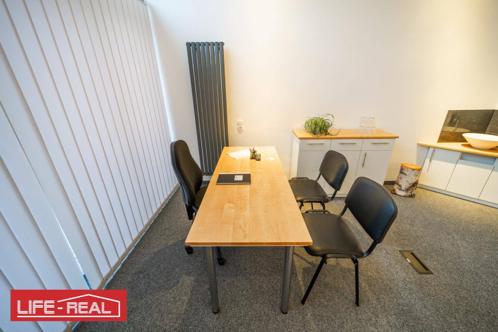 Detailansicht eines Bürobereichs mit Schreibtisch, Stühlen, vertikalen Jalousien und einem modernen Heizkörper.