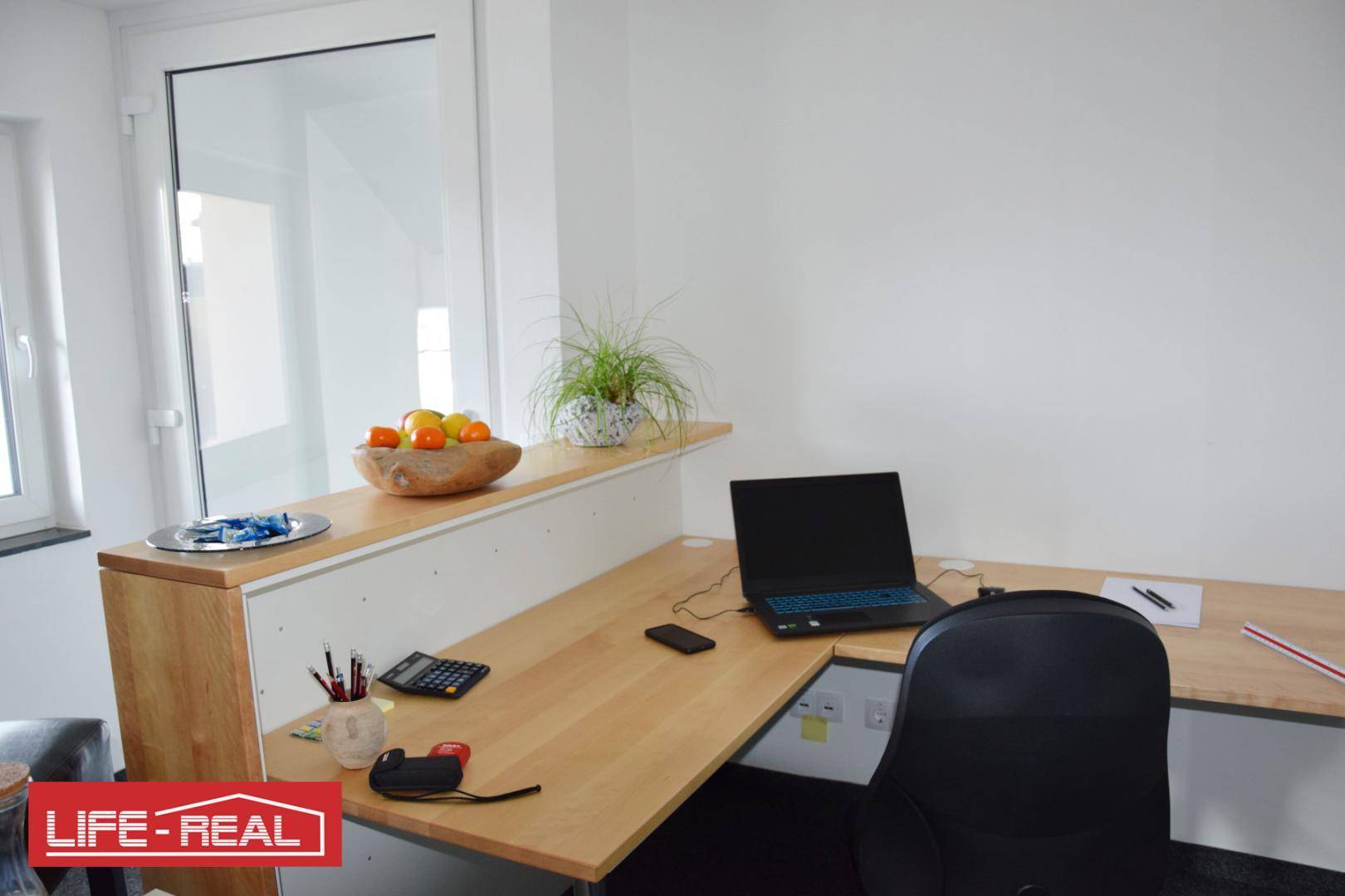 Detailansicht eines hellen Holzschreibtisches mit Laptop, Obstschale und einer Pflanze in einem modernen Büro.