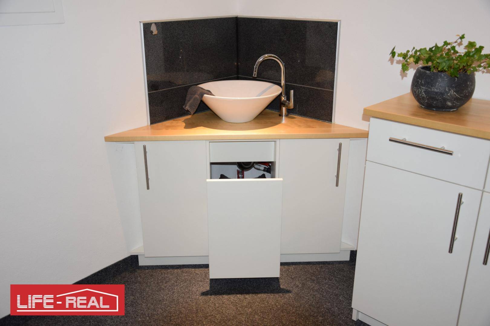 Kleine Eckspüle mit weißem Waschbecken, schwarzem Fliesenspiegel und hellen Unterschränken in einem Funktionsbereich.