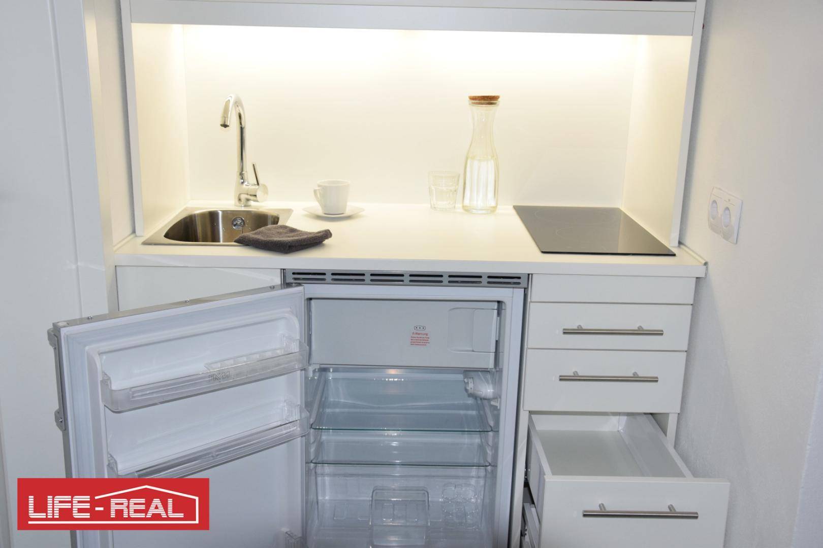 Kompakte Küchenzeile mit Spüle, Induktionskochfeld, Kühlschrank und praktischen Schubladen in Weiß.