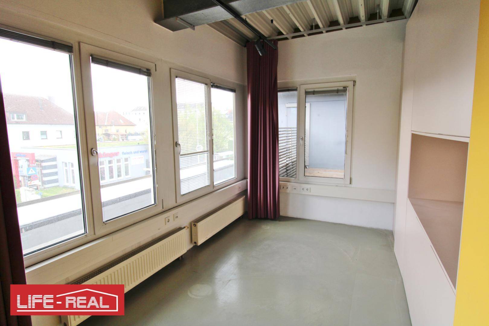 Kleiner Büroraum mit großen Fenstern, Blick auf die Straße und einem Heizkörper.