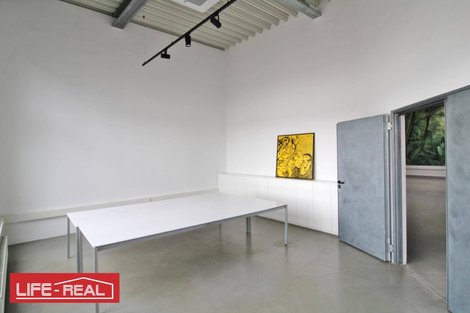 Heller Büroraum mit weißem Tisch, Kunstwerk an der Wand und Zugang zu einem weiteren Raum.