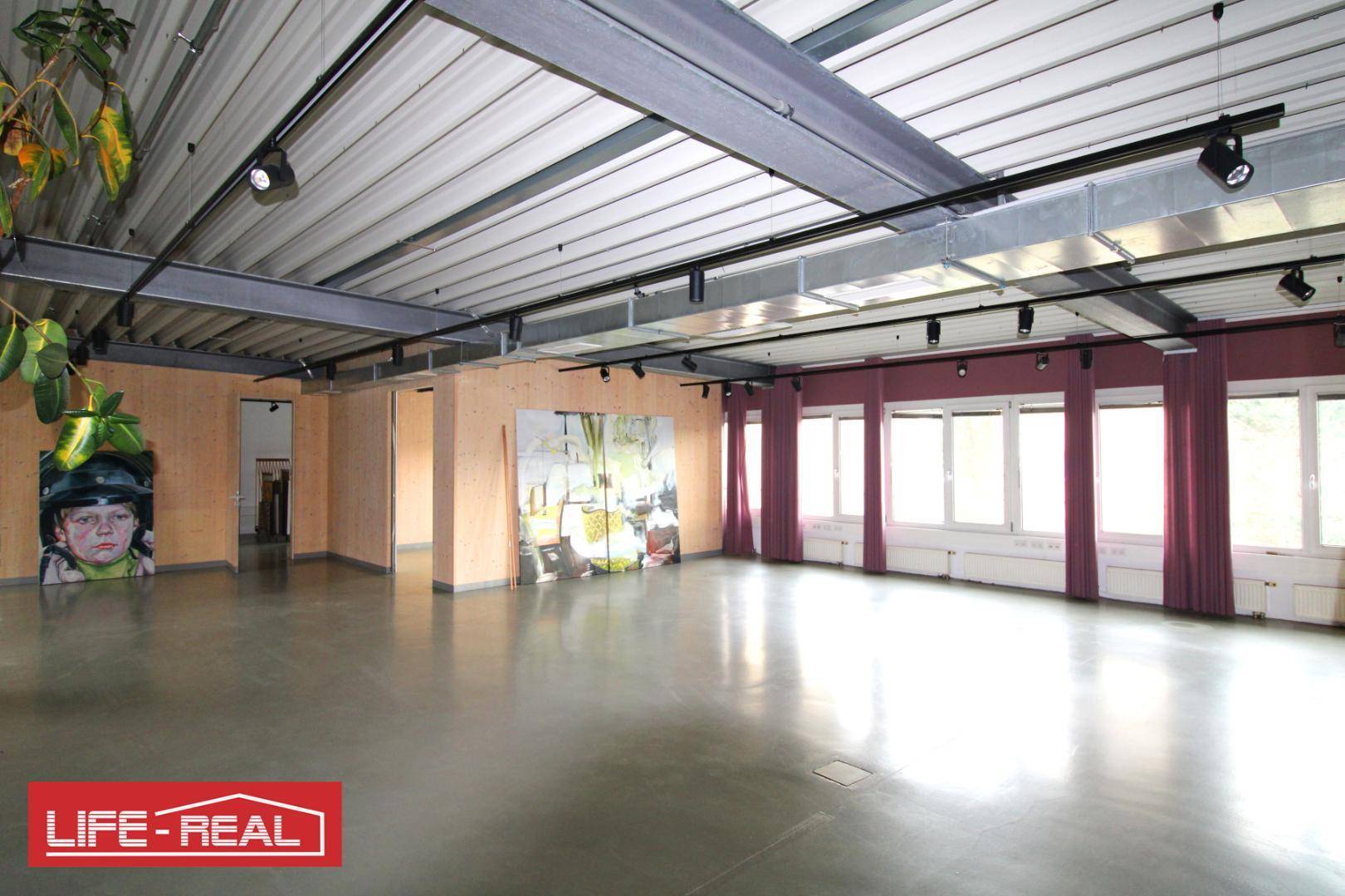 Weitläufiger Gewerberaum mit hohen Decken, Betonboden und einer Fensterfront mit bordeauxroten Vorhängen.