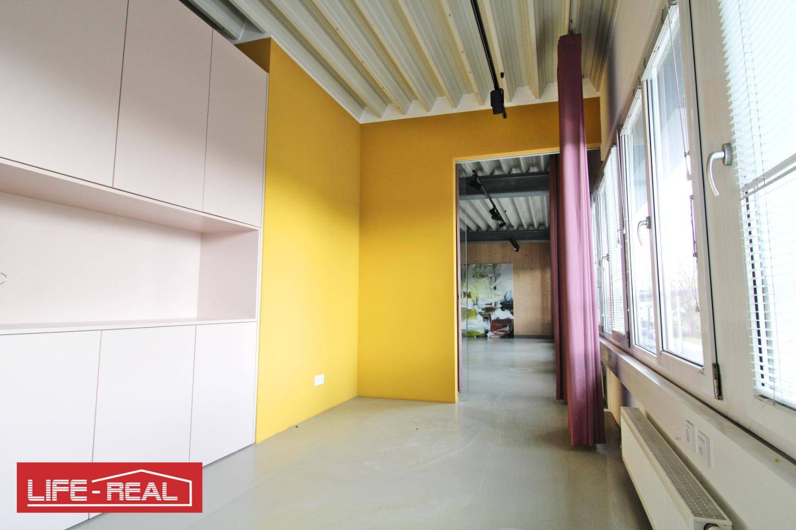 Büroraum mit einer auffälligen gelben Wand, weißen Einbauschränken und großen Fenstern.