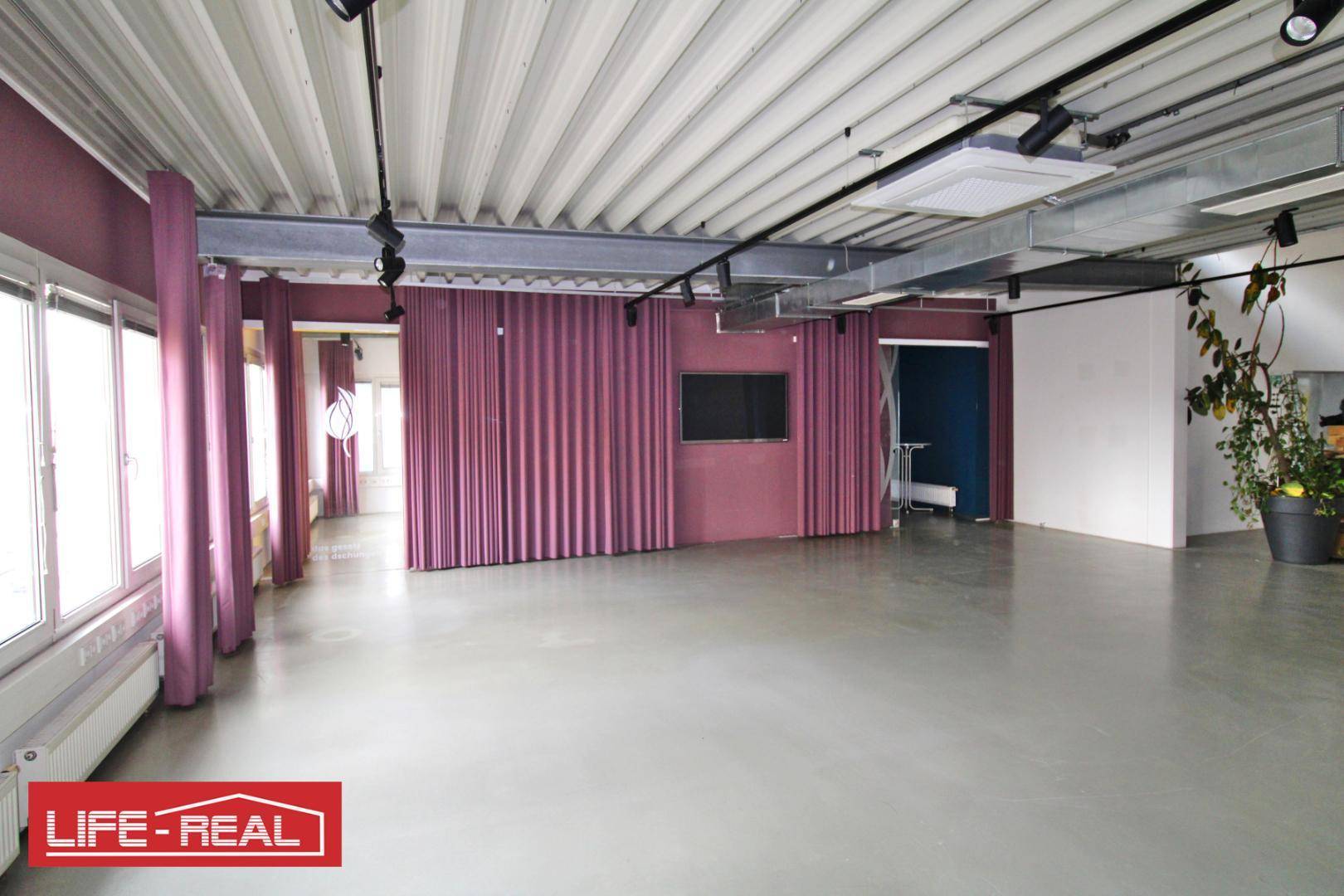 Heller Gewerberaum mit hohen Decken, Betonboden und einer Fensterfront mit bordeauxroten Vorhängen.