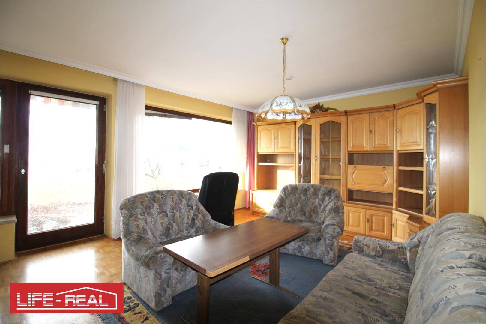 Helles Wohnzimmer mit großen Fenstern, Teppichboden und traditionellen Holzmöbeln.