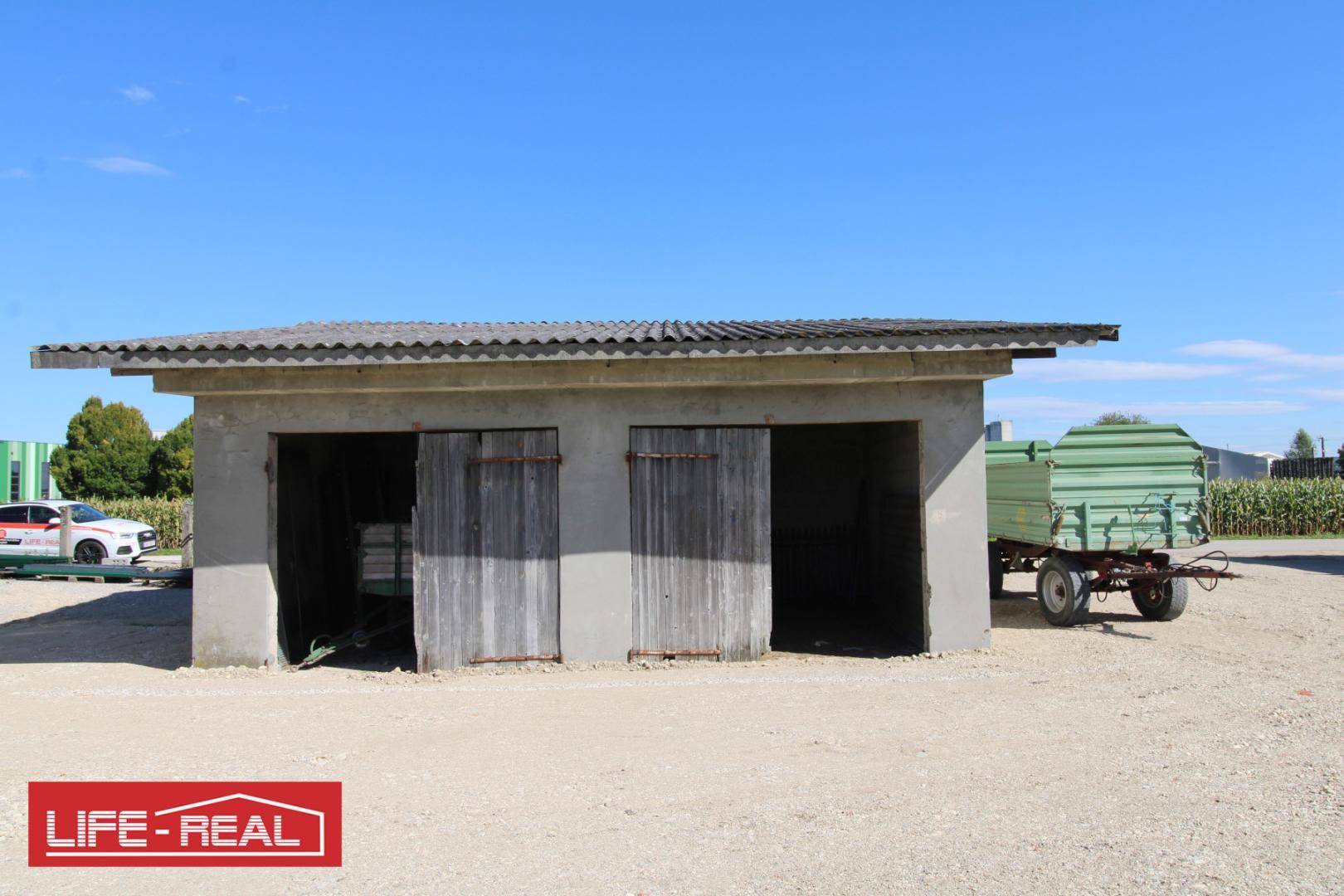 Ein einfaches landwirtschaftliches Gebäude mit zwei Holztüren und einem Anhänger auf einem Schotterplatz.