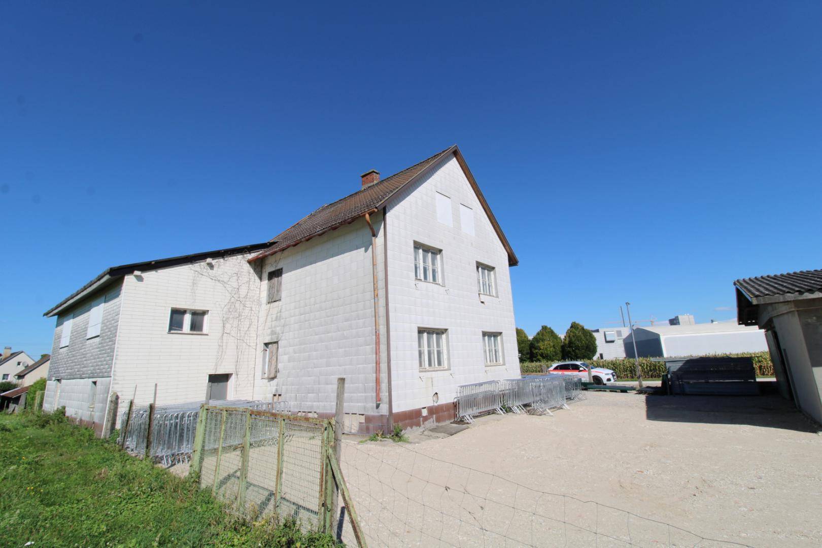 Älteres Wohnhaus mit weißer Fassade und Satteldach, umgeben von einem Schotterplatz und Zäunen.