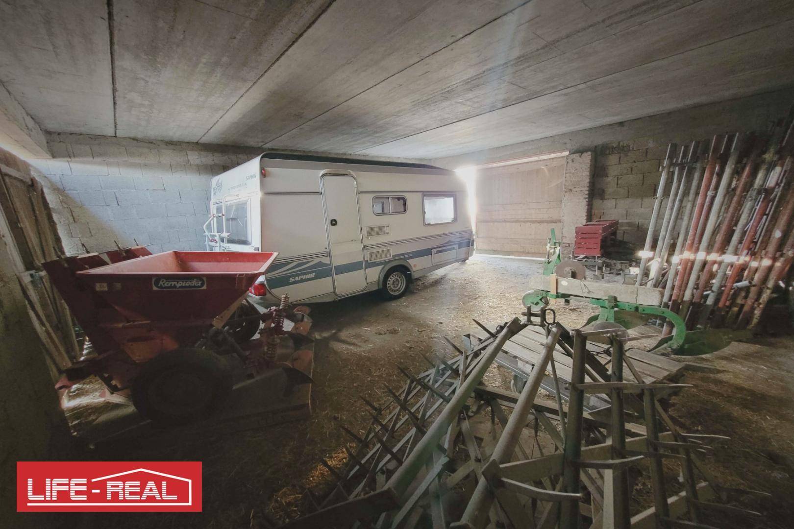 Innenansicht eines Lagerraums mit einem Wohnwagen und landwirtschaftlichen Geräten, beleuchtet durch die Türöffnung.